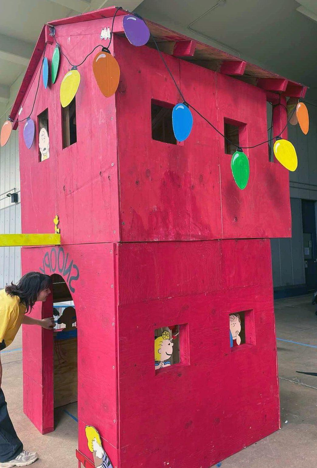 A red, tall, tiny home stands assembled and decorated with two-dimentional holiday lights and Peanuts characters.